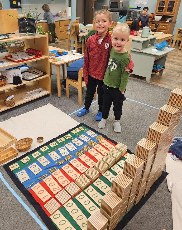 Students smile for a photo next to a large completed project that they worked on together in a Montessori classroom
